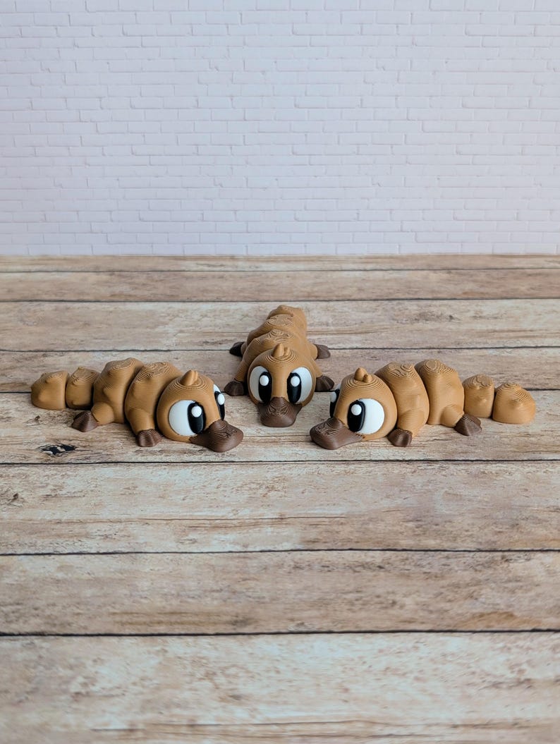 May include: Three brown platypus figurines with large eyes and duck-like bills are arranged on a wooden surface. The figurines are lying down, with their bodies segmented. The background features a white brick wall.