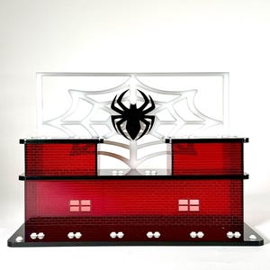 May include: A red brick-patterned display stand with a black spider emblem and web design. The stand has a rectangular base and two levels, with a clear panel featuring a spider web and a black spider.