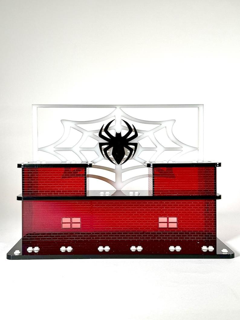 May include: A red brick-patterned display stand with a black spider emblem and web design. The stand has a rectangular base and two levels, with a clear panel featuring a spider web and a black spider.