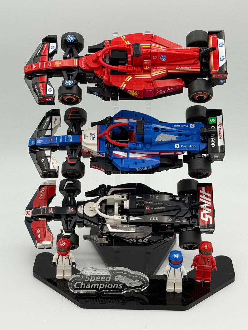 May include: Three Lego Speed Champions race cars on a black display stand. The cars are a red Ferrari, a blue and white car, and a black and red car. Each car has a driver figure next to it.