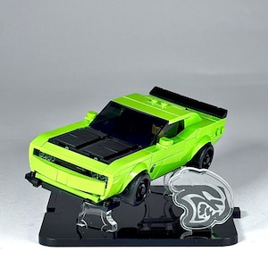 Puede incluir: Un coche de juguete verde lima con detalles en negro, incluyendo un capó y alerón negros. El coche está expuesto sobre un soporte negro, con un emblema de acrílico transparente de un logotipo estilizado. El coche de juguete es un modelo de coche deportivo.