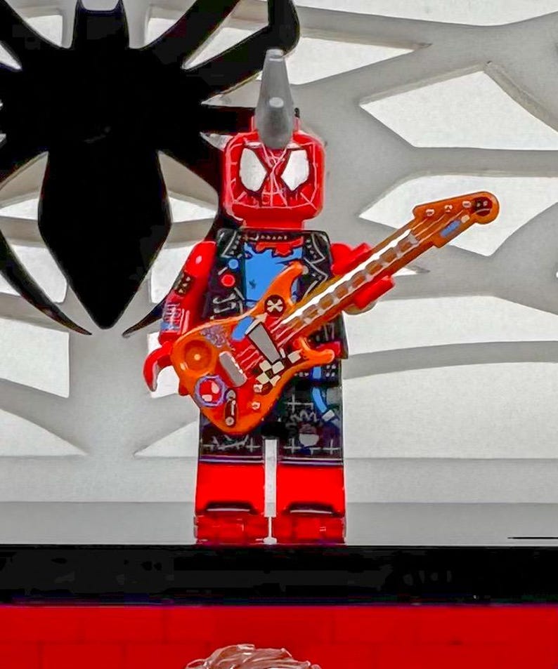 May include: A red and black Lego figure with a mohawk and a guitar. The figure is wearing a black vest, blue shirt, and black shorts. The guitar is orange and brown with a checkered design.