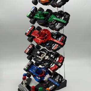 May include: A display stand with five miniature Formula 1 race cars. The cars are in different colors and have different sponsors. The stand is made of clear acrylic and has a black base. The base has the words "Speed Champions" printed on it. There are four miniature figures standing in front of the stand.