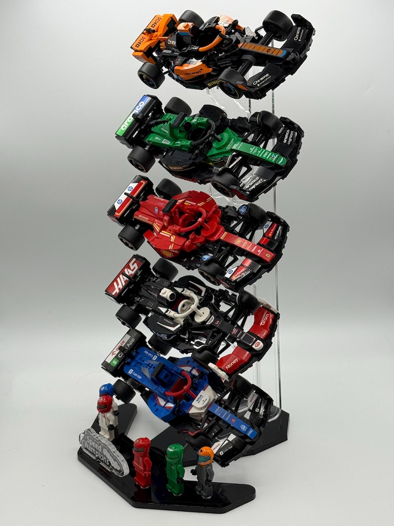 May include: A display stand with five miniature Formula 1 race cars. The cars are in different colors and have different sponsors. The stand is made of clear acrylic and has a black base. The base has the words "Speed Champions" printed on it. There are four miniature figures standing in front of the stand.