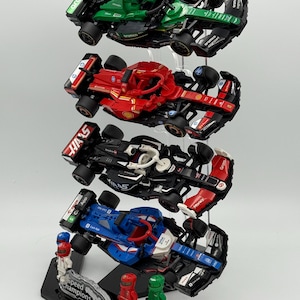 May include: A display stand with four Lego Speed Champions race cars. The cars are stacked on top of each other and are in different colors, including green, red, black, and blue. The stand is made of clear acrylic and has a black base with the words "Speed Champions" printed on it.