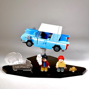 May include: A Lego set featuring a light blue car with a white roof, and three mini-figures. The set includes a white owl, a boy with black hair, and a boy with red hair. A brown rat is also included. The set is displayed on a black stand.