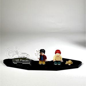 May include: A black display base with three Lego figures and a small animal figure. The Lego figures are of a boy with a red and black plaid shirt, a boy with a yellow shirt, and a white owl. The display base has the text "-76424" and "165 Pieces".