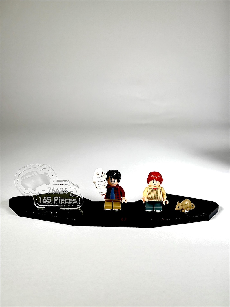 May include: A black display base with three Lego figures and a small animal figure. The Lego figures are of a boy with a red and black plaid shirt, a boy with a yellow shirt, and a white owl. The display base has the text "-76424" and "165 Pieces".
