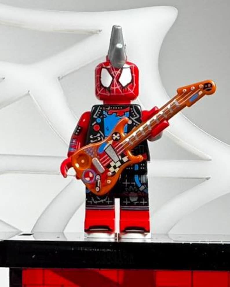 May include: A red and black Lego Spider-Man figure with a silver mohawk and a guitar. The guitar is orange and decorated with various designs. The figure is standing on a black surface with a white web design in the background.