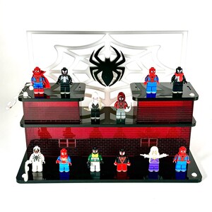 May include: A multi-tiered display stand featuring various Spider-Man character figurines. The stand has a red brick design with a white backdrop displaying a spider web and spider logo. The figurines are in various colors, including red, blue, black, and white.