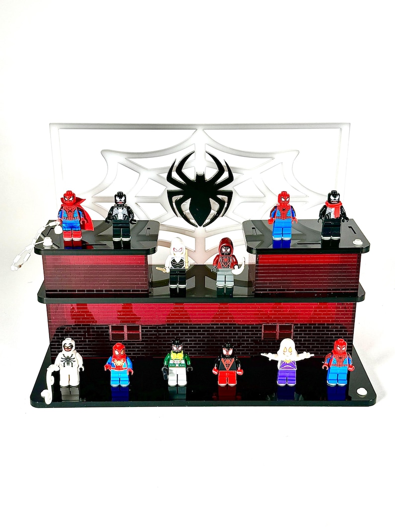 May include: A multi-tiered display stand featuring various Spider-Man character figurines. The stand has a red brick design with a white backdrop displaying a spider web and spider logo. The figurines are in various colors, including red, blue, black, and white.