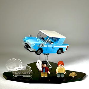 May include: A Lego set featuring a light blue flying car with a white roof, and miniature figures of Harry Potter, Ron Weasley, and Hedwig the owl. The set includes a black base with the text "76424 165 Pieces".