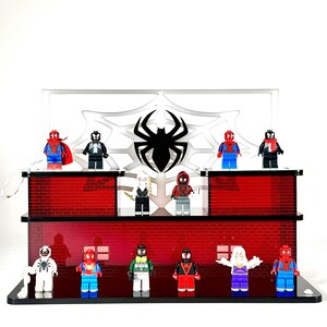 May include: A display featuring various Spider-Man-themed Lego minifigures. The display has a red brick facade and a white spiderweb design with a black spider emblem. The minifigures are in various colors and costumes.