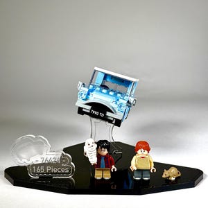 May include: A light blue Lego car model with the license plate "7990 TD" is suspended above a black display base. The base also holds Lego figures of Harry Potter, Ron Weasley, Hedwig the owl, and a small brown rat. A clear sign reads "76424 165 Pieces."