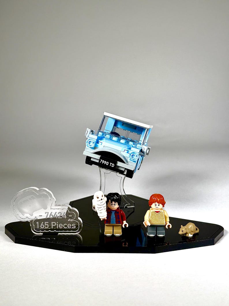 May include: A light blue Lego car model with the license plate "7990 TD" is suspended above a black display base. The base also holds Lego figures of Harry Potter, Ron Weasley, Hedwig the owl, and a small brown rat. A clear sign reads "76424 165 Pieces."