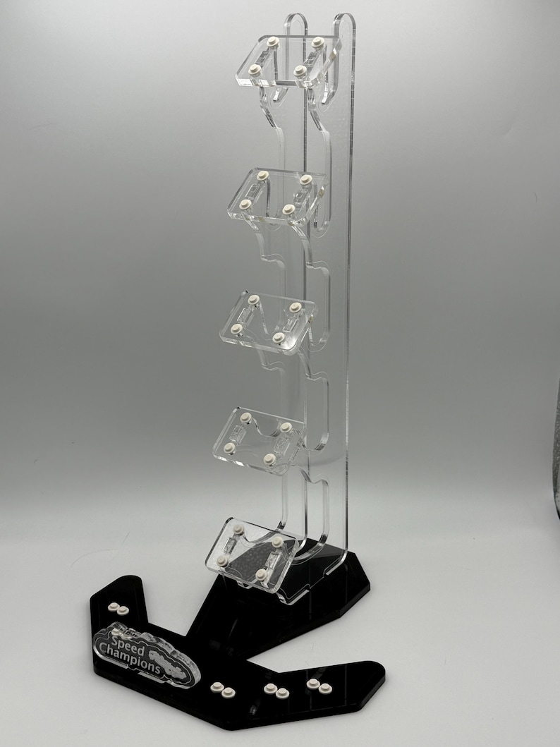 May include: A clear acrylic display stand with five slots for holding small cars. The stand has a black base with the words "Speed Champions" printed on it.