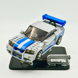 May include: A gray and blue toy car with a blue spoiler and a white and blue stripe down the center. The car is on a black base with a clear sign that reads "NISSAN SKYLINE R".