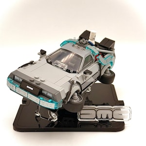 May include: A gray and black DeLorean Time Machine model, with blue accents, is displayed on a black stand. The model has the letters "DMC" on the front and on the stand. The car appears to be hovering.