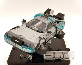 May include: A gray and black DeLorean Time Machine model, with blue accents, is displayed on a black stand. The model has the letters "DMC" on the front and on the stand. The car appears to be hovering.