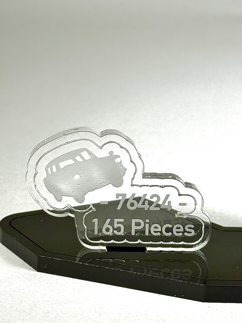 May include: A clear acrylic car-shaped ornament with the number "76424" and the text "1.65 Pieces" etched into it. The ornament sits on a black base and is set against a light gray background.