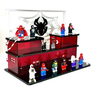 May include: A display featuring multiple Spider-Man action figures on a two-tiered red brick-style platform. A white spiderweb and a black spider silhouette are in the background. The figures are in various costumes.