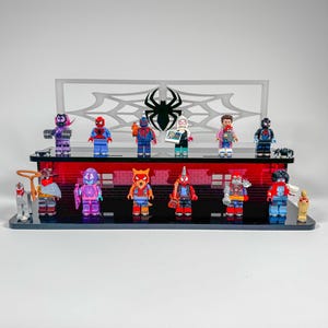 Può includere: Una mensola espositiva con statuette a tema supereroi. La mensola ha una base nera con un accento rosso e un retro in acrilico trasparente con un design a ragnatela e un logo a ragno. Le statuette sono di vari colori e design.