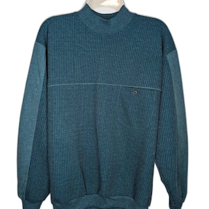 Peut inclure: Pull en tricot côtelé bleu sarcelle avec un col montant et des manches longues. Les manches ont un panneau bleu sarcelle contrastant. Un détail de ligne horizontale traverse la poitrine, avec un petit accent rond rouge. Le pull est suspendu à un cintre en plastique transparent.