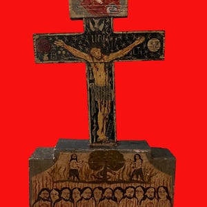 May include: A hand-painted wooden crucifix with a red background. The cross features a depiction of Jesus Christ, with additional painted figures and symbols. The base of the cross has a painted scene with multiple figures. The artwork is done in a folk art style.