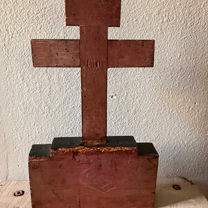 May include: A rustic, wooden cross painted in shades of red and brown. The cross is constructed from several pieces of wood and has a rectangular base. The wood shows signs of age and wear, adding to its vintage appeal. The cross is displayed against a textured white wall.