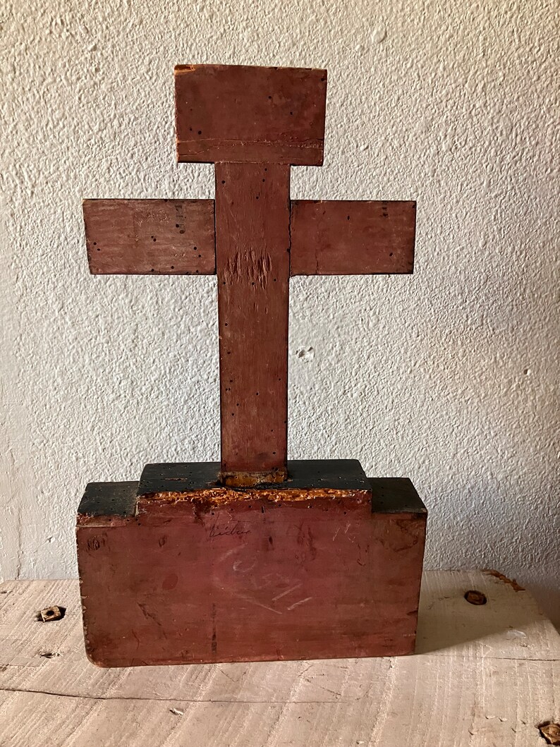 May include: A rustic, wooden cross painted in shades of red and brown. The cross is constructed from several pieces of wood and has a rectangular base. The wood shows signs of age and wear, adding to its vintage appeal. The cross is displayed against a textured white wall.