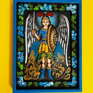 May include: Hand-painted religious icon of an angel with a sword and shield, set in a blue wooden frame. The angel wears a blue and yellow tunic, with white wings and a red feathered helmet. The frame is decorated with blue flowers and gold accents.