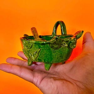 Op de afbeelding: Een kleine, groene keramische kom met een handvat en drie voetjes, vastgehouden in een hand. De kom heeft een textuur met bladachtige details en bevat een klein houten voorwerp. De achtergrond is effen oranje.