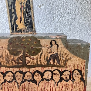May include: A folk art painting on wood depicting a biblical scene. The artwork features figures, a tree with apples, and a serpent. The color palette includes shades of brown, black, and red. The painting is mounted on a wooden structure.