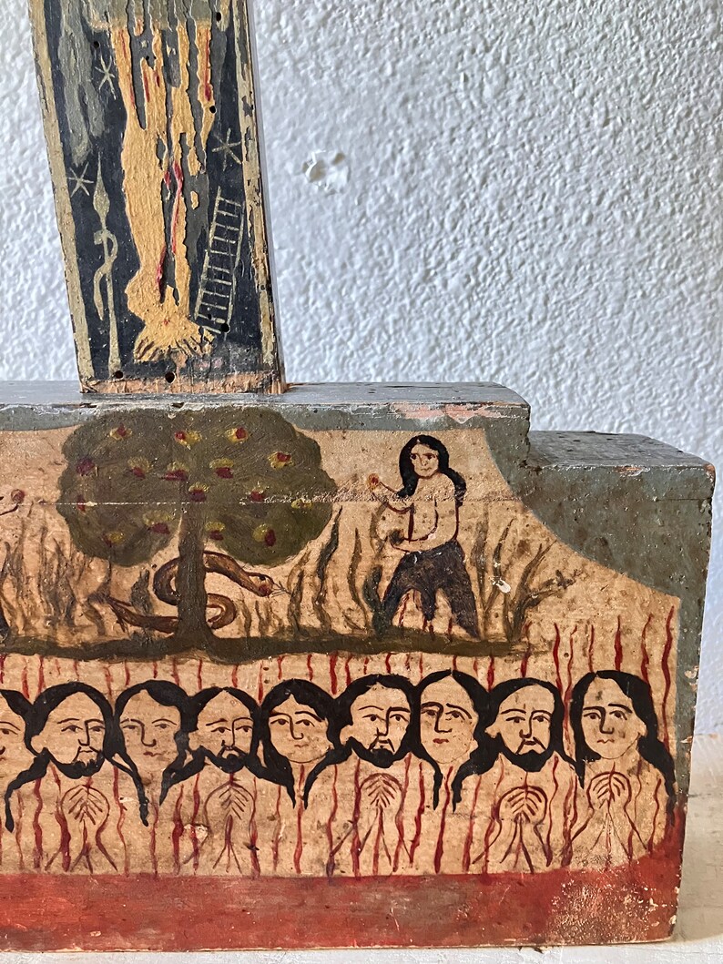May include: A folk art painting on wood depicting a biblical scene. The artwork features figures, a tree with apples, and a serpent. The color palette includes shades of brown, black, and red. The painting is mounted on a wooden structure.