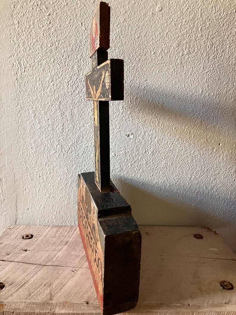 May include: A tall, wooden religious artifact with a cross design. The piece is painted black, red, and beige, with detailed illustrations. The cross is mounted on a rectangular base, and the overall style suggests a vintage or antique aesthetic.