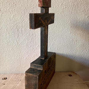 May include: A hand-carved wooden cross with a black base and crossbar. The cross features painted religious imagery in red, brown, and beige tones. The base has a detailed relief carving. The piece has an aged, rustic appearance.