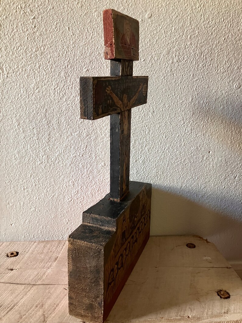 May include: A hand-carved wooden cross with a black base and crossbar. The cross features painted religious imagery in red, brown, and beige tones. The base has a detailed relief carving. The piece has an aged, rustic appearance.