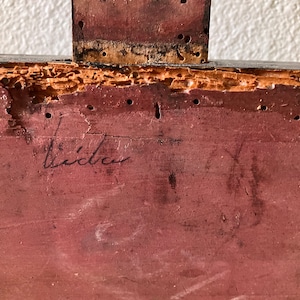 May include: Close-up of a section of a wooden object, possibly furniture. The wood is painted a reddish-brown color and shows signs of age and wear, including small holes and a signature that reads "Lida".