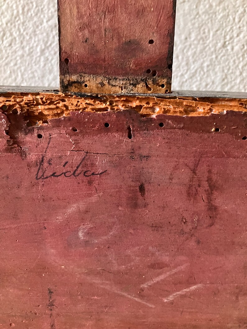 May include: Close-up of a section of a wooden object, possibly furniture. The wood is painted a reddish-brown color and shows signs of age and wear, including small holes and a signature that reads "Lida".