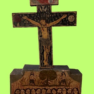 May include: A hand-painted wooden crucifix with a detailed depiction of Jesus Christ on the cross. The cross features painted figures, stars, and the letters INRI. The base shows figures and a tree. The artwork is on a green background.