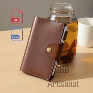 以下が含まれることがあります： 真鍮のボタン留めが付いた茶色の革製ジャーナル。ジャーナルは閉じられており、ステッチされた縁があります。画像には、黄色の液体が入ったガラス瓶、白いマグカップ、およびテキスト「JSB Artisanat」も含まれています。
