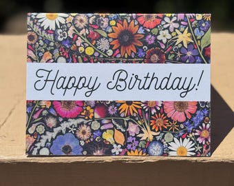 Op de afbeelding: Een rechthoekige verjaardagskaart met een bloemenontwerp. De kaart heeft een levendige collage van kleurrijke bloemen tegen een donkere achtergrond. Een witte banner over het midden toont de woorden "Happy Birthday!" in elegant schrift.