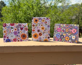 Op de afbeelding: Drie bloemen wenskaarten met kleurrijke bloemenontwerpen. De kaarten bevatten een verscheidenheid aan bloemen, waaronder zonnebloemen, madeliefjes en andere wilde bloemen. De kaarten worden weergegeven op een houten oppervlak tegen een achtergrond van groen gebladerte.