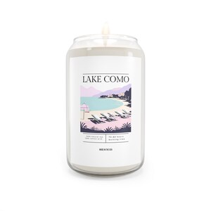 May include: A white candle in a glass jar shaped like a can. The candle has a label that says "LAKE COMO" and features a graphic of a lake with mountains and beach chairs. The label also says "100% natural soy, 100% cotton wick, 70-90 hours burning time, Made in the USA."