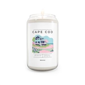 May include: A white candle in a glass jar shaped like a can. The label says "Cape Cod" and features a cartoon illustration of a blue house with a pink roof and a pink flower garden. The label also says "100% natural soy, 100% cotton wick, 50-60 hours burning time, Made in the USA."