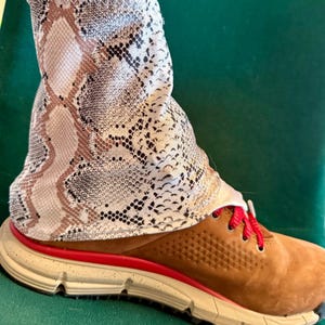 May include: A brown suede shoe with red laces and a red sole trim. The shoe is paired with a fabric bootie featuring a snakeskin pattern in shades of brown, beige, and black. The shoe has a cream-colored sole.