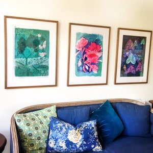 May include: Three framed art prints hanging on a white wall above a blue couch with pillows. The left print features a green tree with white birds flying around it. The middle print features a pink flower with green leaves on a blue background. The right print features a dark background with colorful butterflies.