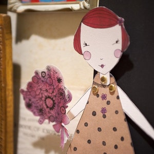 May include: Paper doll with red hair, wearing a brown dress with white polka dots and a gold necklace. The doll is holding a pink and purple bouquet of flowers.