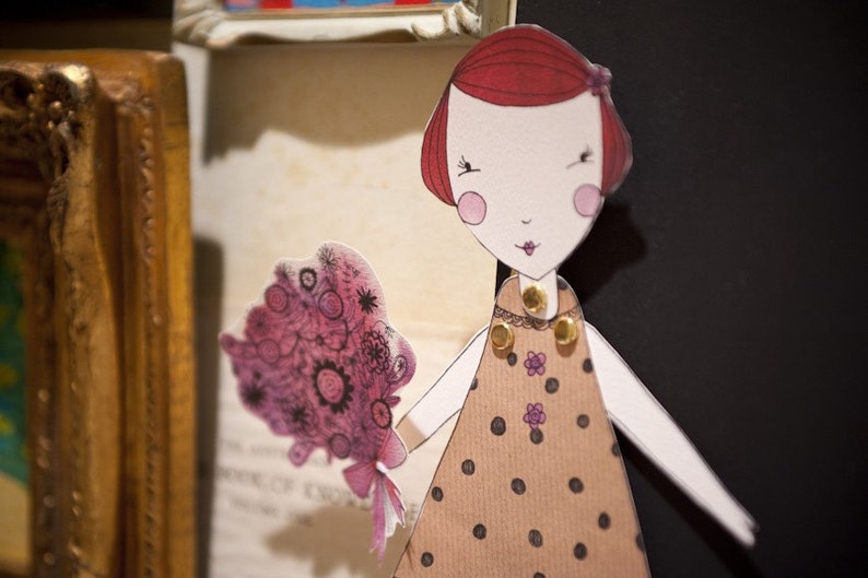 May include: Paper doll with red hair, wearing a brown dress with white polka dots and a gold necklace. The doll is holding a pink and purple bouquet of flowers.