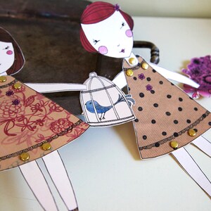 May include: Two paper dolls with brown and black polka dot dresses, one holding a birdcage with a blue bird inside. The dolls have red and brown hair and are wearing gold buttons.